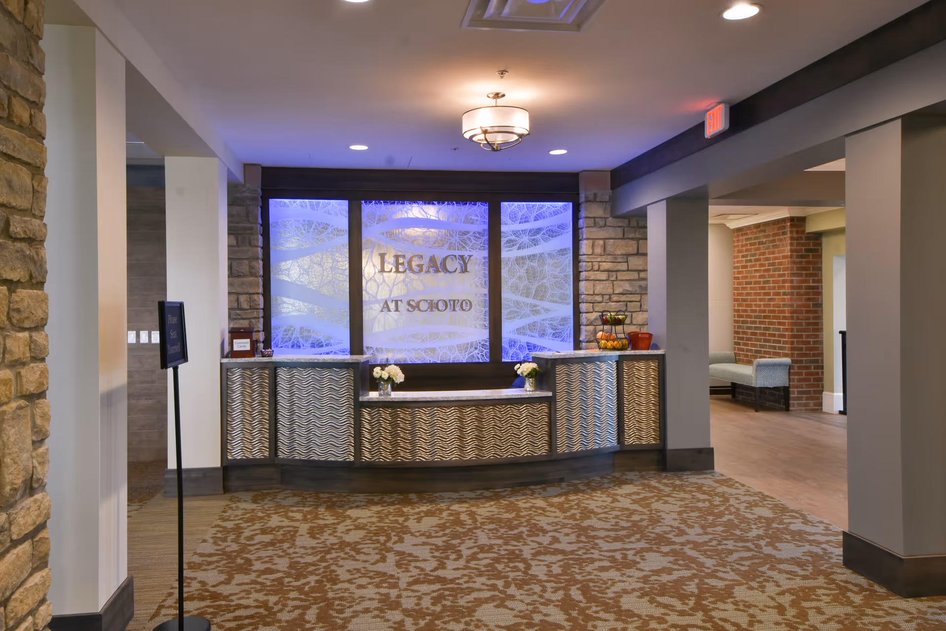 Reception area with a decorative front desk featuring a wavy pattern, illuminated backdrop with the text 'LEGACY AT SCIOTO', stone and brick walls, and a carpeted floor with a floral pattern. There are flowers on the desk and a fruit basket on the right side. The area is well-lit with ceiling lights.