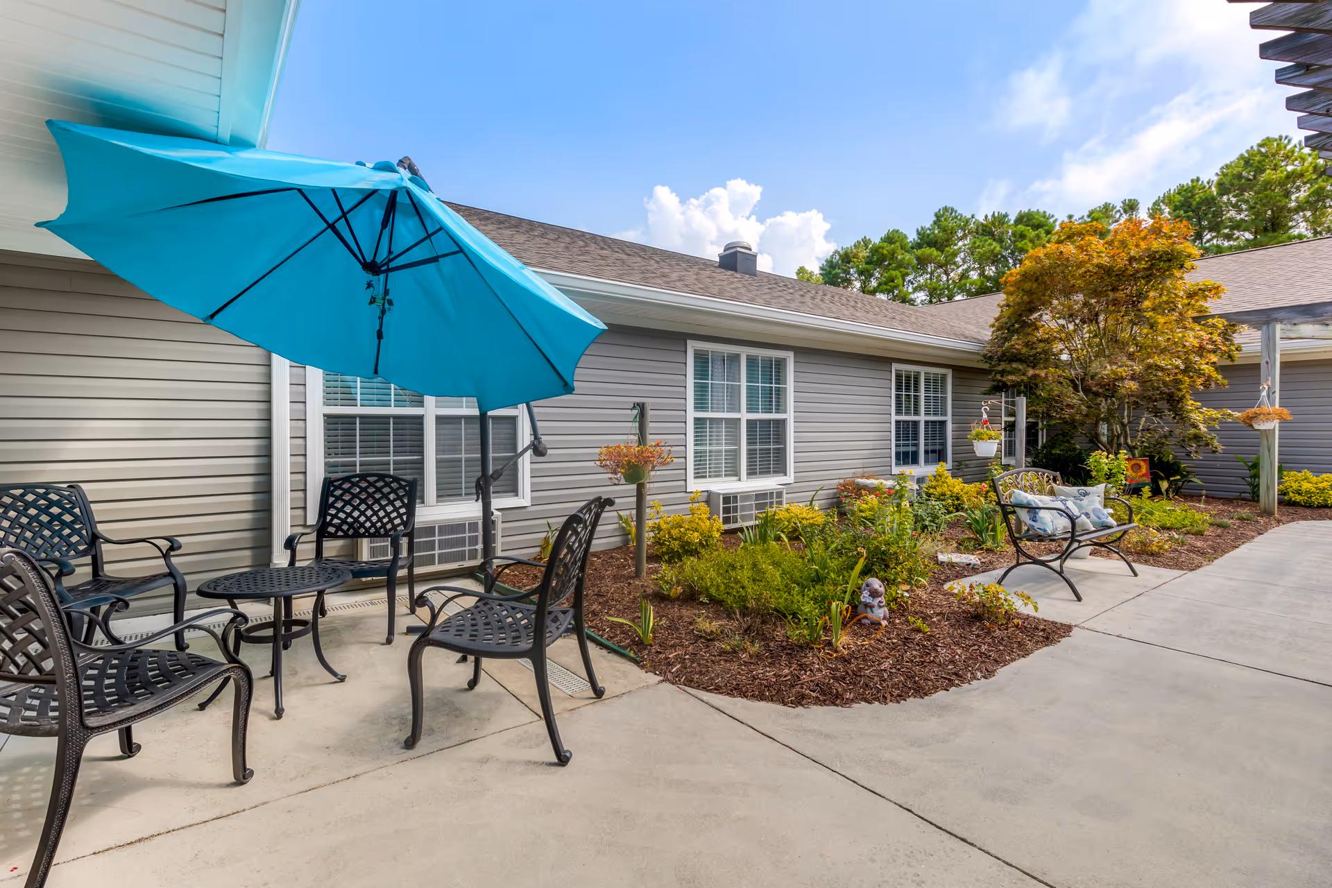 Outdoor patio area at Brookdale Morehead City featuring a round metal table with four matching chairs and a large blue umbrella. The patio is adjacent to a garden with various plants and a small tree, and there is a bench with decorative pillows along the walkway. The building exterior is gray with white-trimmed windows under a clear blue sky.