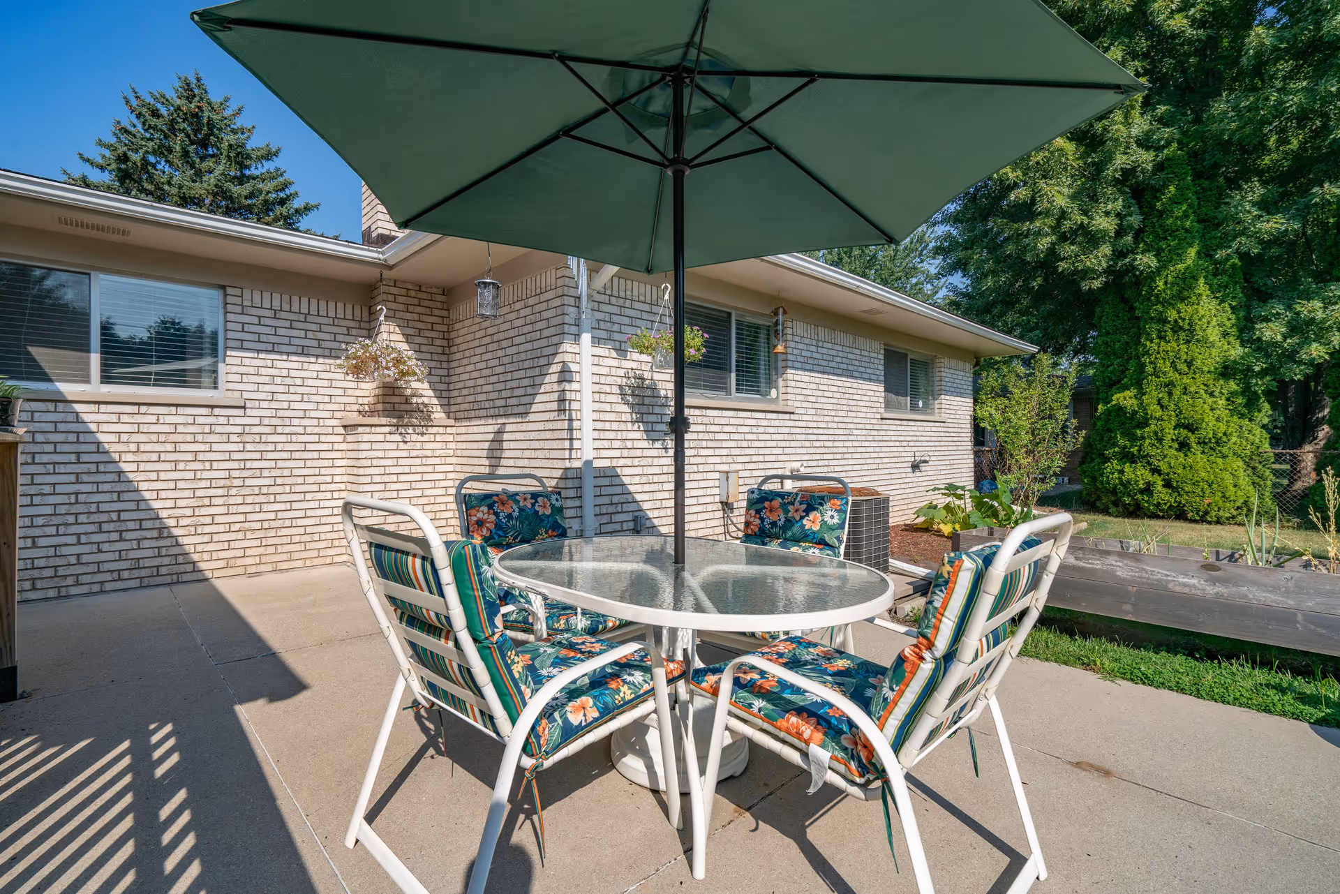 Outdoor patio area with a round glass table and four cushioned chairs with floral and striped patterns. A large green umbrella is open above the table. The patio is adjacent to a light-colored brick building with windows and hanging flower pots. Trees and greenery are visible in the background under a clear blue sky.