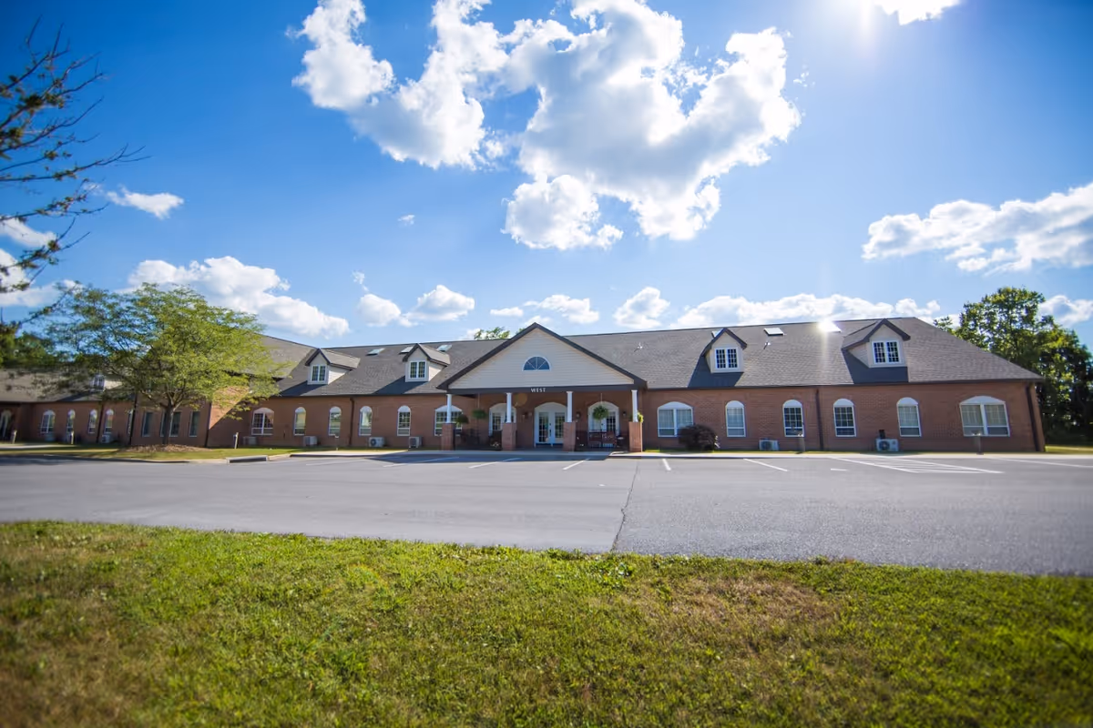 Exterior view of a single-story brick building with multiple windows and dormers under a bright blue sky with scattered clouds. The building has a covered entrance with columns and a parking lot in front, surrounded by green grass and trees.
