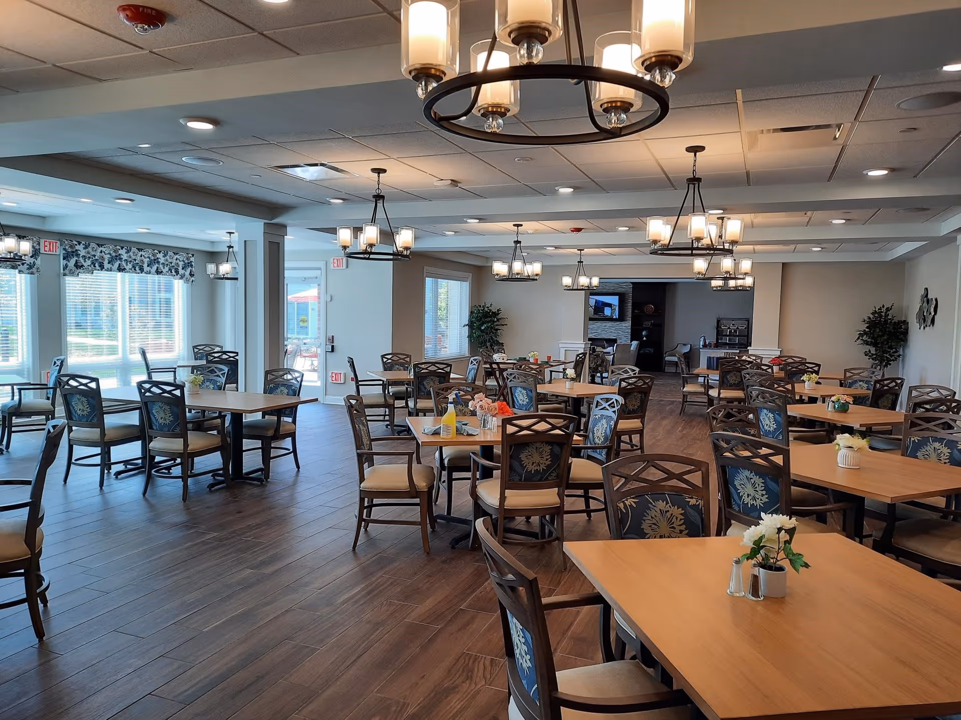 A spacious dining room with multiple wooden tables and cushioned chairs arranged neatly. The room has large windows with floral valances allowing natural light to fill the space. Several chandeliers hang from the ceiling providing warm lighting. There are small flower arrangements on the tables and a few potted plants near the walls.