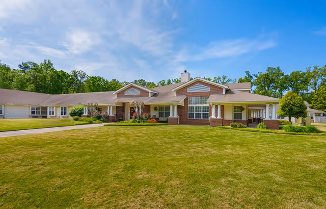 Front exterior of a single-story brick senior living building with columned covered porches, large windows, and a wide grassy lawn under a blue sky.