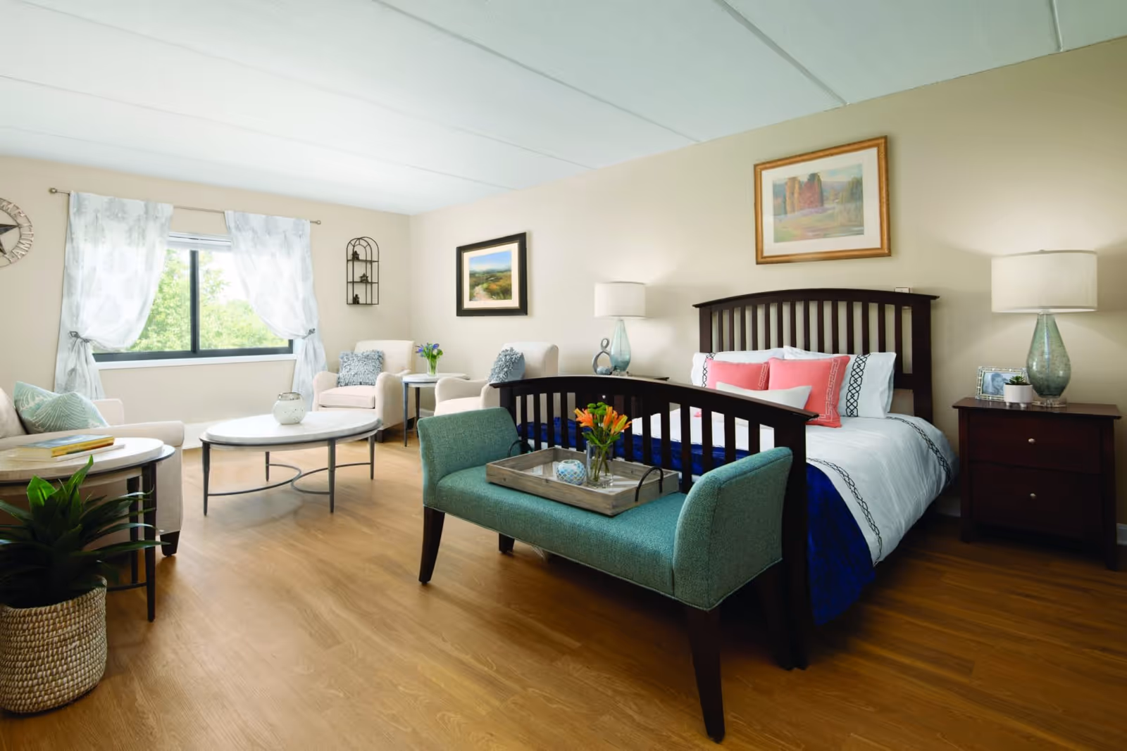 A bright and cozy bedroom with a wooden bed featuring white and pink bedding. At the foot of the bed is a teal upholstered bench with a tray holding a vase of orange flowers and a decorative item. To the right of the bed is a dark wooden nightstand with a lamp, a framed photo, and a small plant. The room has light-colored walls with two framed paintings and a window with white patterned curtains letting in natural light. There are two beige armchairs with blue pillows and a round coffee table in front of them. A green plant in a woven basket is placed on the wooden floor near the armchairs.