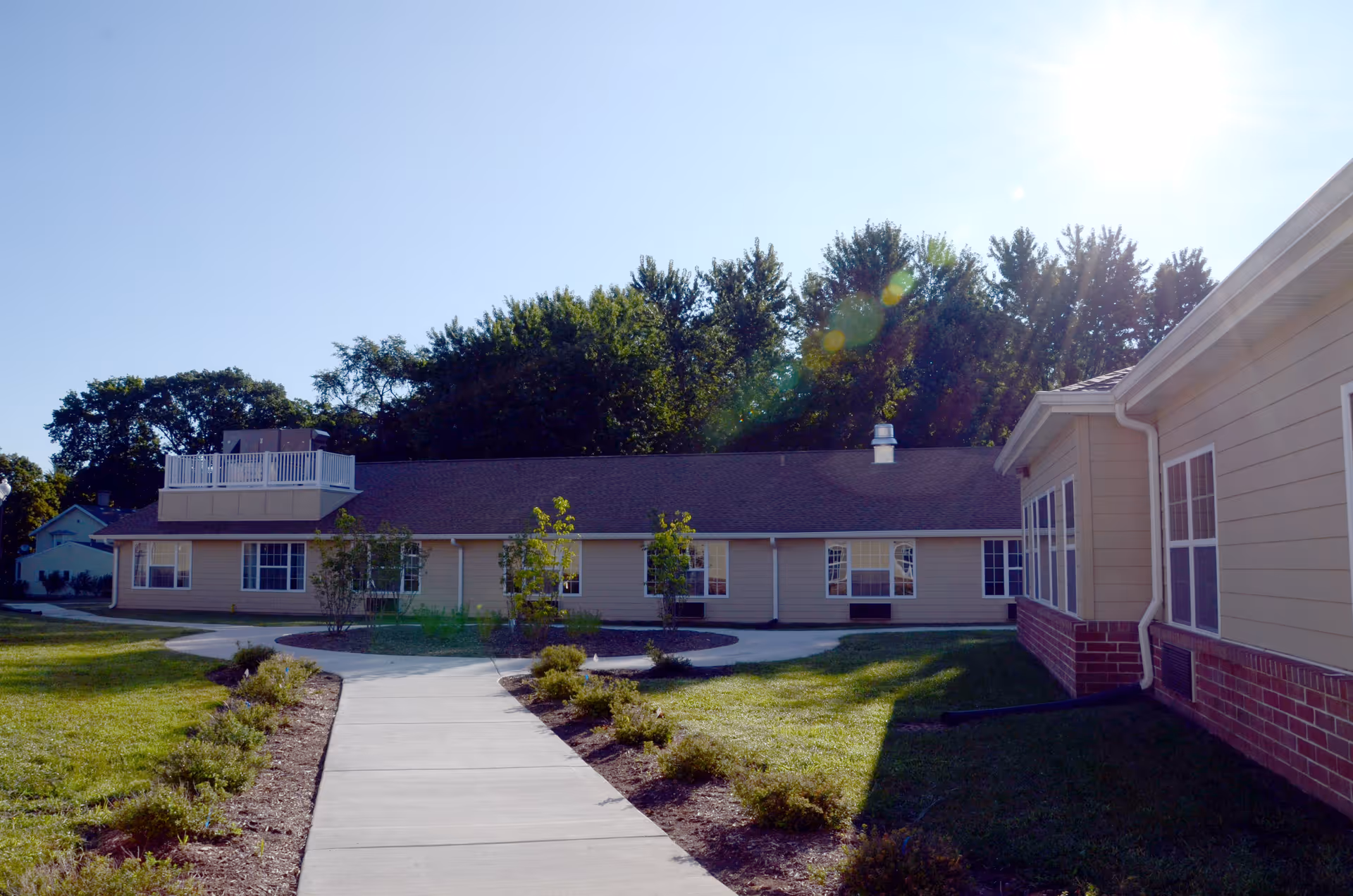 Exterior view of Pavilion of Ottawa facility showing a single-story building with beige siding and a brick base, surrounded by a well-maintained lawn and small shrubs along a concrete walkway, with trees in the background under a clear blue sky.
