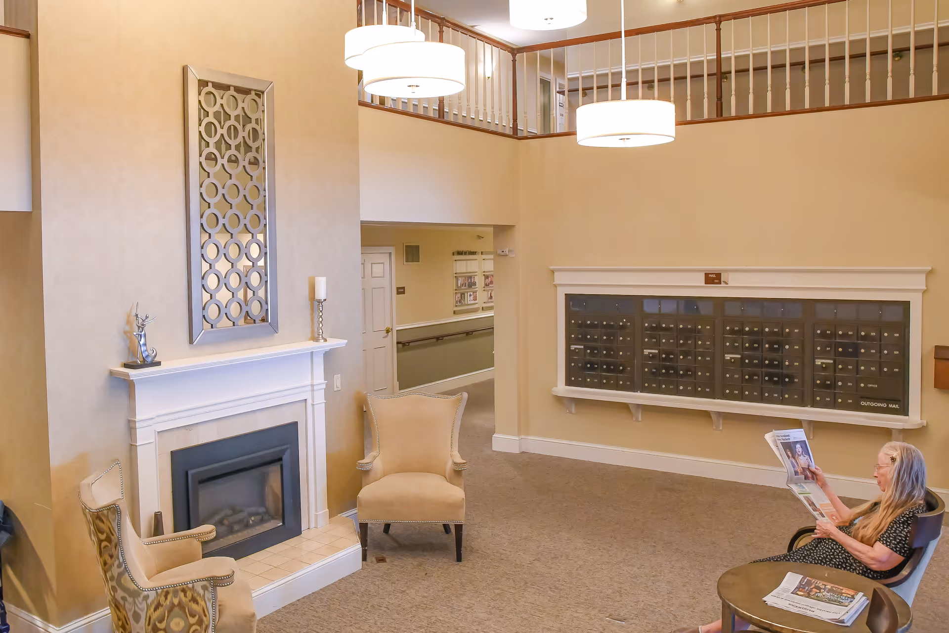 A cozy senior living facility common area with beige walls and carpet. There is a white fireplace with decorative items on the mantel, two beige armchairs, and a woman sitting in a chair reading a newspaper near a small round table. On the far wall, there is a large set of mailboxes labeled 'Outgoing Mail'. The area has a high ceiling with two hanging light fixtures and a wooden railing on the upper level.