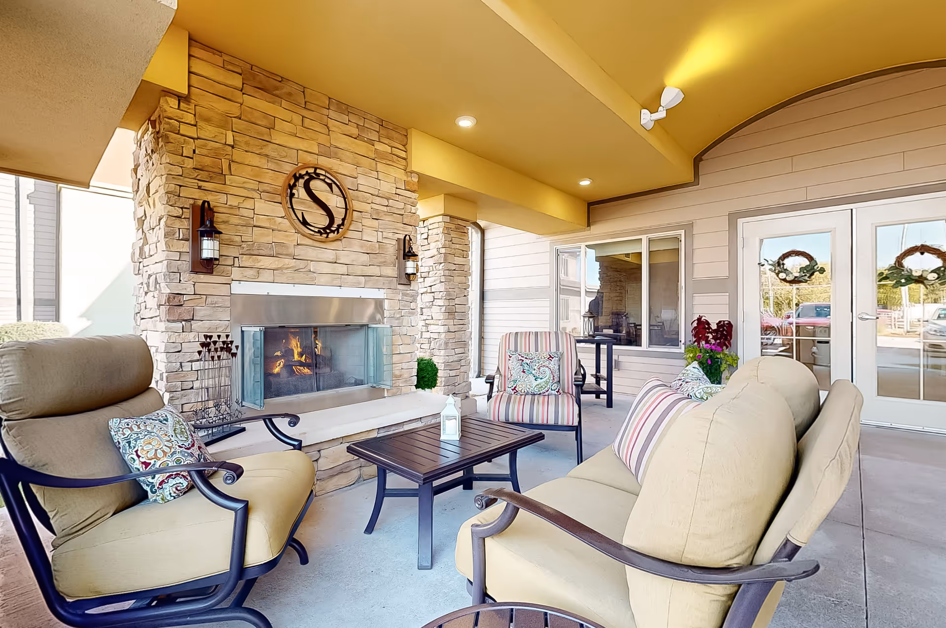 Outdoor covered patio area with cushioned seating including armchairs and a loveseat arranged around a small wooden table. A stone fireplace with a metal 'S' emblem above it is lit, providing warmth. The area is decorated with colorful pillows, potted plants, and has double glass doors with wreaths leading inside.