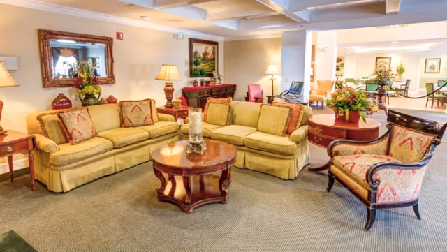 A cozy living room area in a senior living facility featuring two yellow sofas with patterned cushions, a wooden round coffee table with candles, an upholstered armchair with a red and gold pattern, side tables with lamps, floral arrangements, a large mirror on the wall, and framed artwork. The room has a carpeted floor and is well-lit with ceiling lights.