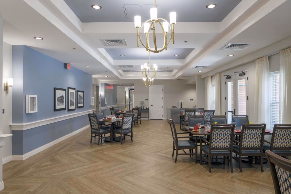 A spacious dining room with multiple tables and chairs arranged neatly. The room features light wood flooring, blue and white walls, and modern chandeliers hanging from a coffered ceiling. The tables are set with red cups, plates, and napkins. Large windows with white curtains allow natural light into the room.