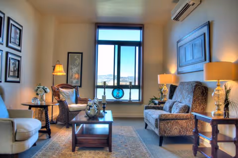 A cozy living room in a senior living facility featuring a large window with a view, two patterned armchairs, a beige armchair, a wooden coffee table with decorative items, side tables with lamps, framed artwork on the walls, and a carpeted floor.
