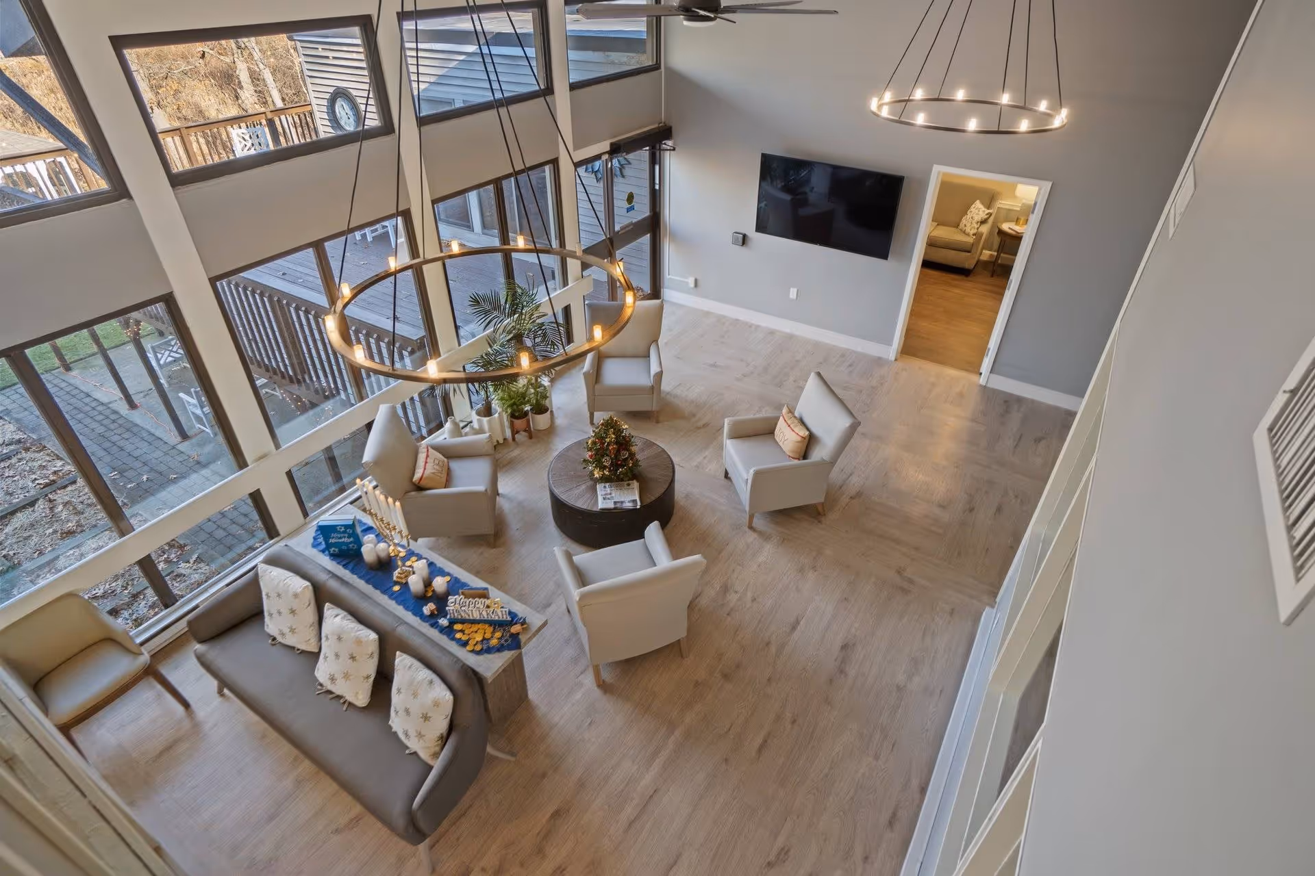 A spacious living room with large floor-to-ceiling windows allowing natural light to fill the space. The room features a modern circular chandelier, a round coffee table with a floral centerpiece, four armchairs arranged around the table, and a sofa with decorative pillows. A flat-screen TV is mounted on a gray wall, and a doorway leads to another room with a beige armchair and side table. Outside, a wooden deck and patio area are visible through the windows.