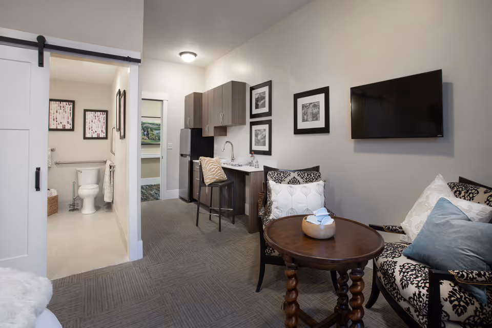 Interior view of a senior living residence featuring a small living area with patterned armchairs, a round wooden table with a decorative bowl, a wall-mounted flat screen TV, and a kitchenette with cabinets, a sink, and a bar stool. To the left, a sliding barn door opens to a bathroom with a toilet, grab bars, and framed artwork on the walls.