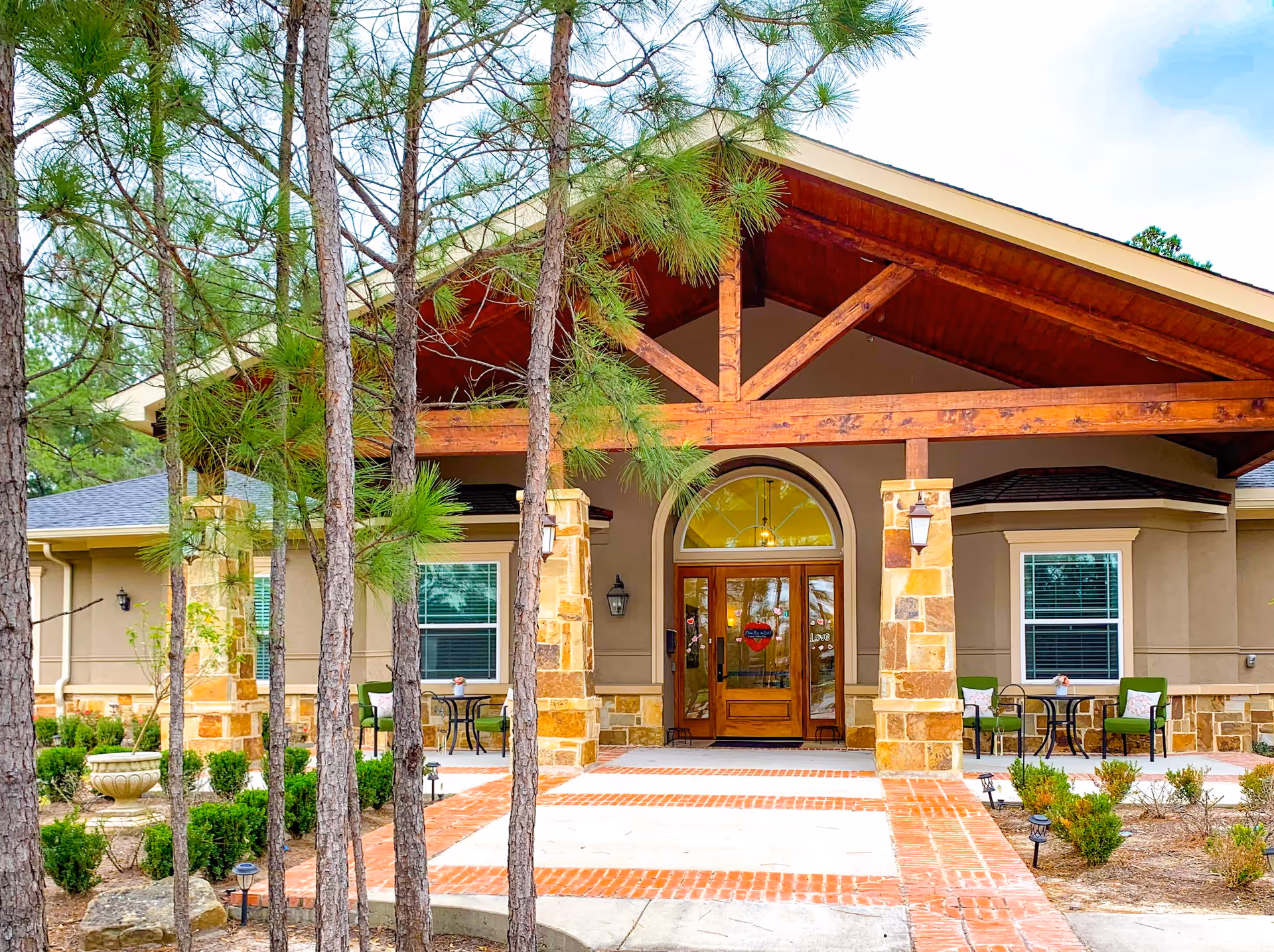Front exterior view of Village Green Memory Care Community Cypress featuring a covered entrance with wooden beams, stone pillars, glass double doors, and outdoor seating with green chairs and small tables. There are trees and landscaped bushes in the foreground.