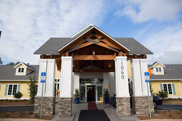 Entrance of a senior living facility with a covered porch supported by stone and white pillars, a peaked roof with wooden beams, and the number 1060 displayed on one pillar. The building is yellow with white trim and has windows with green shutters. There are two handicap parking signs visible near the entrance and some potted plants and chairs under the porch.