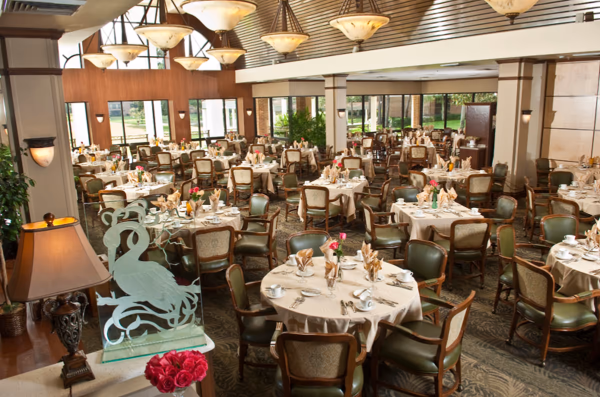 Large dining room with many round tables set with tableware and chairs beneath hanging lights.