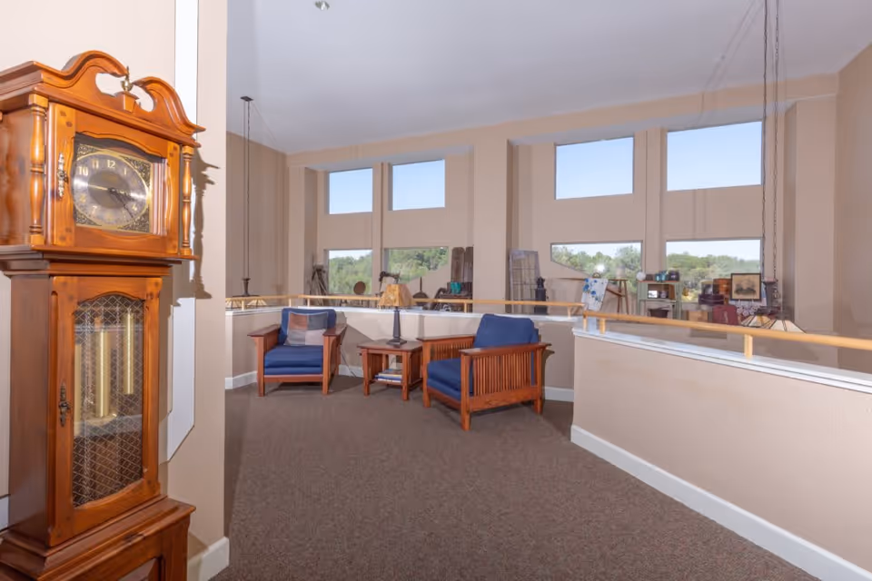 A cozy sitting area in a senior living facility with two wooden armchairs featuring blue cushions, a wooden side table with a lamp, and a large wooden grandfather clock. The space has beige walls, carpeted floors, and large windows letting in natural light with a view of greenery outside.