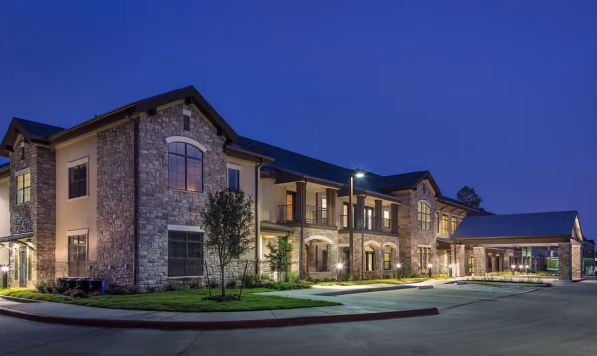 Exterior view of a two-story senior living facility building at dusk with stone and stucco facade, illuminated windows, a covered entrance, and a well-maintained driveway and landscaping.