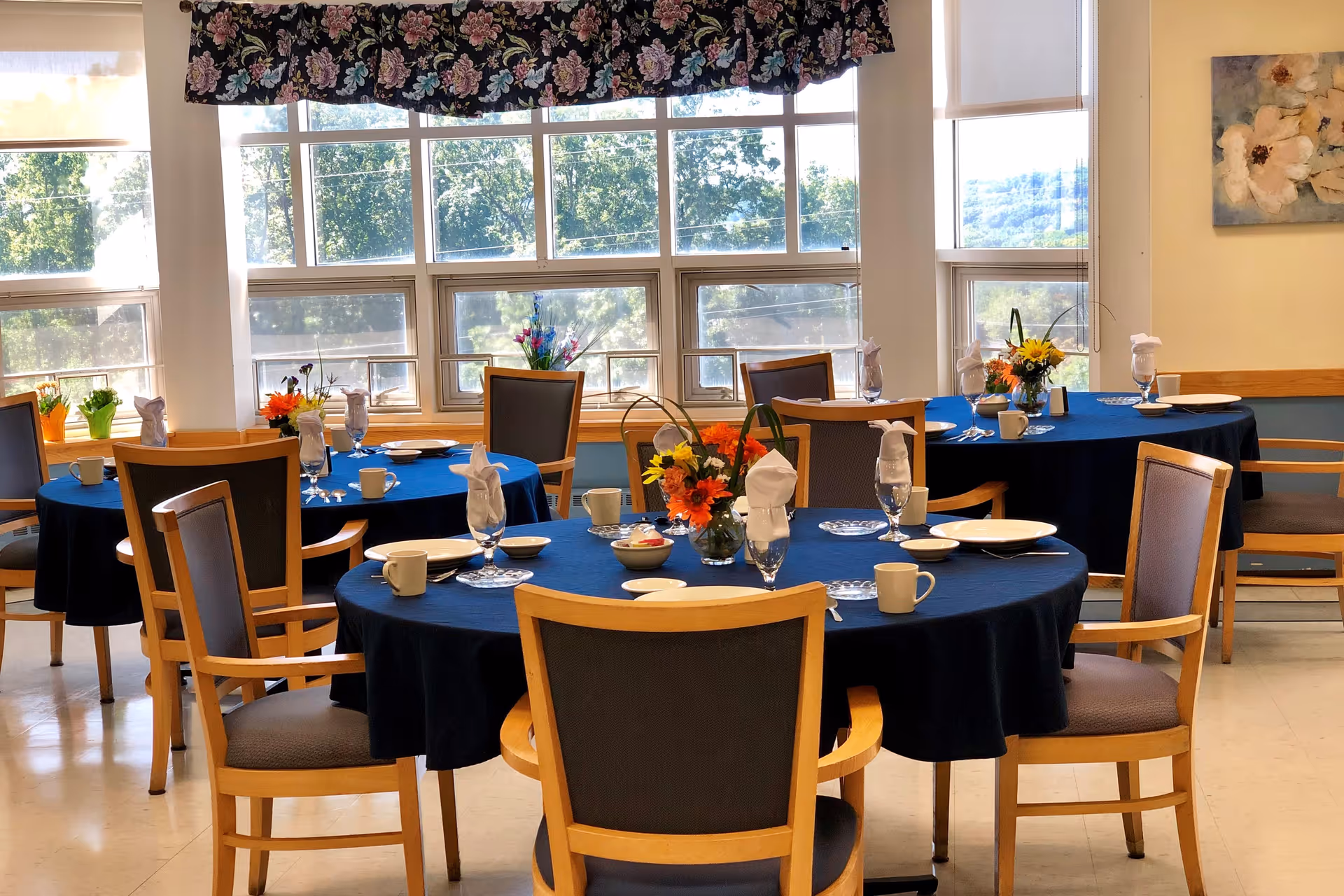 A bright dining room with round tables covered in dark blue tablecloths, each set with white plates, cups, napkins folded in glasses, and floral centerpieces. Large windows with floral valances let in natural light and show greenery outside.