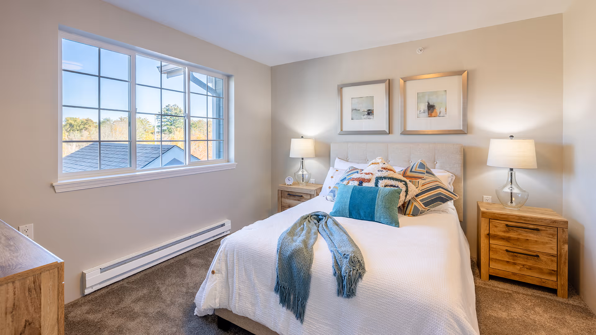 A cozy bedroom with a large window letting in natural light. The bed is neatly made with white bedding, multiple decorative pillows, and a blue throw blanket. There are two wooden nightstands on either side of the bed, each with a lamp. Two framed artworks hang above the bed. The room has beige walls and carpeted flooring.