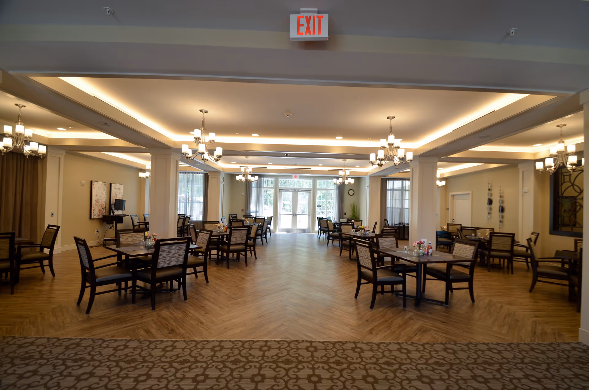 A spacious dining room with multiple tables and chairs arranged neatly. The room features wooden flooring, chandeliers hanging from the ceiling, and large windows at the far end letting in natural light. The walls are painted in a light color, and there is an exit sign above the doorway at the back.
