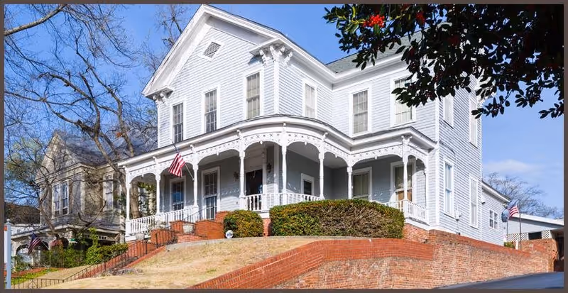A large, white, two-story historic house with a wraparound porch featuring decorative trim. The house is elevated on a brick foundation with a sloped lawn and brick retaining walls. American flags are displayed on the porch and near the side of the house. Leafless trees and a clear blue sky are visible in the background.