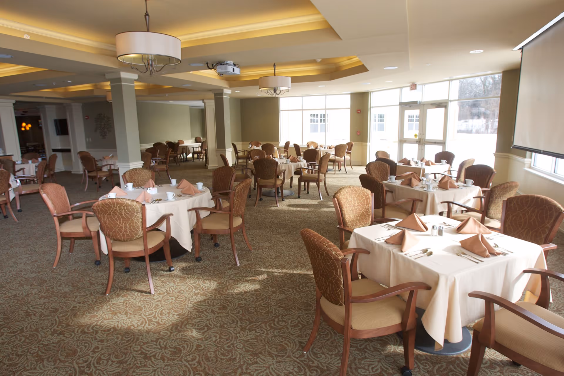 A spacious dining room with multiple round and square tables covered with white tablecloths, each set with folded beige napkins, cups, and silverware. The room has large windows letting in natural light, beige walls, carpeted floor with a patterned design, and ceiling lights with modern fixtures.