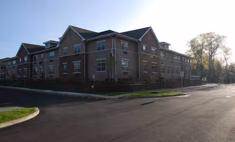 Brick senior living facility viewed from parking lot with landscaped grounds and clear blue sky.
