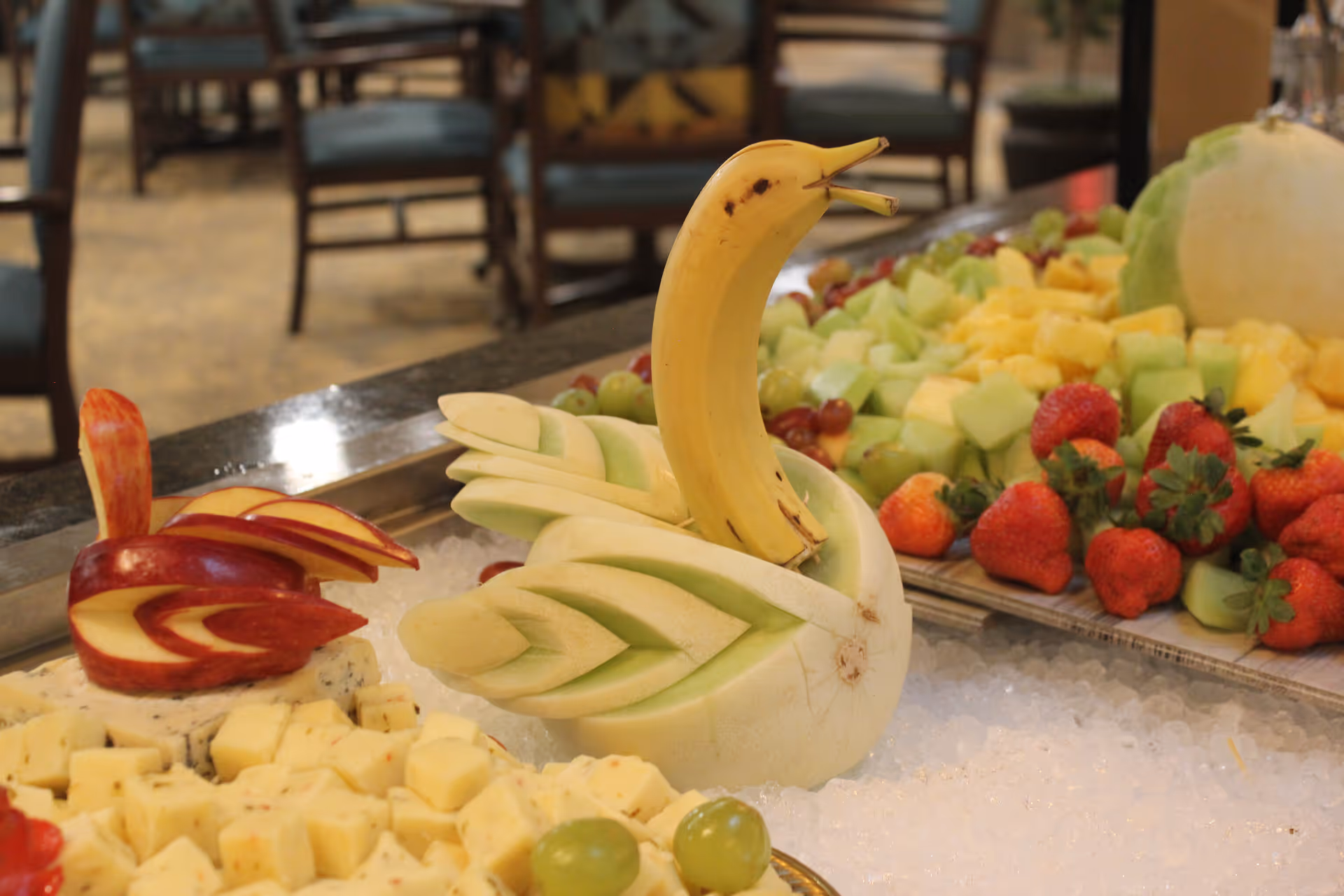 A close-up of a fruit and cheese display featuring a carved melon shaped like a swan with a banana as its neck and head, alongside a red apple carved into a bird shape. Surrounding the carvings are cubes of cheese, grapes, strawberries, and other cut fruits on a bed of ice, with chairs and tables blurred in the background.