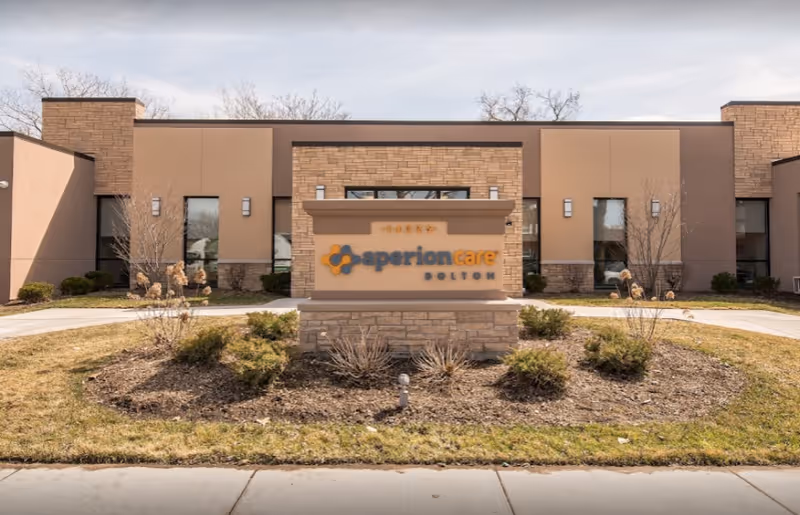 Front exterior view of Aperion Care Dolton building with a stone and beige facade, a sign displaying the facility name, and landscaped bushes and grass in front.