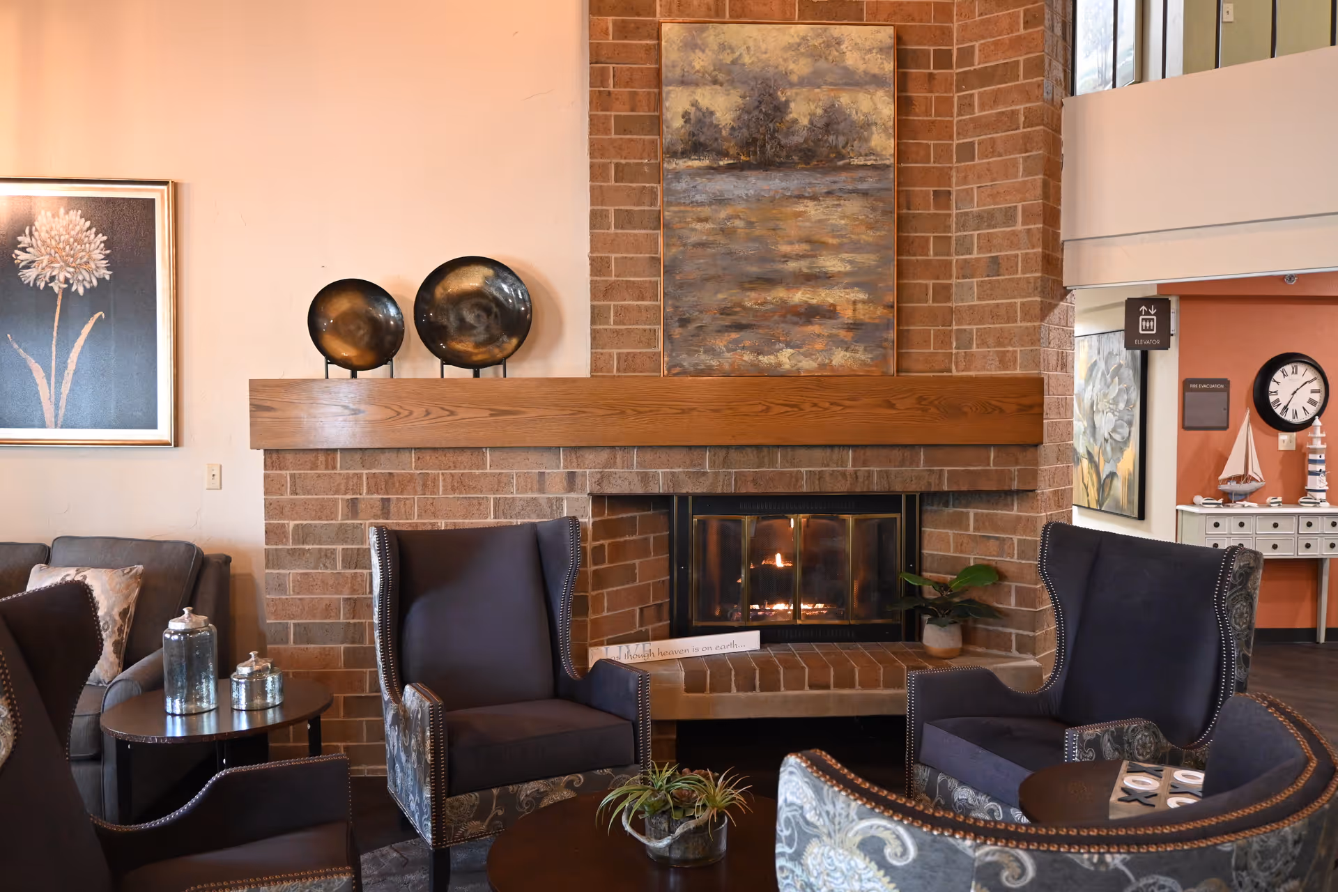 Cozy seating area with upholstered armchairs arranged around a lit brick fireplace and decorative artwork.