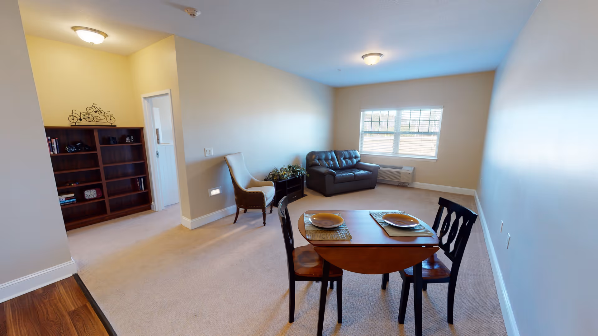 Furnished living room with a small dining table and chairs, a sofa and armchair, bookshelf, and a window letting in natural light.