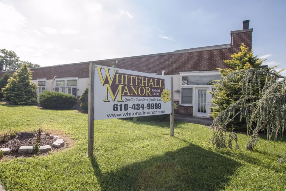 Exterior view of Whitehall Manor assisted living facility showing a brick building with windows and a door, a green lawn with bushes and trees, and a large sign in the foreground displaying the facility name, phone number, and website.