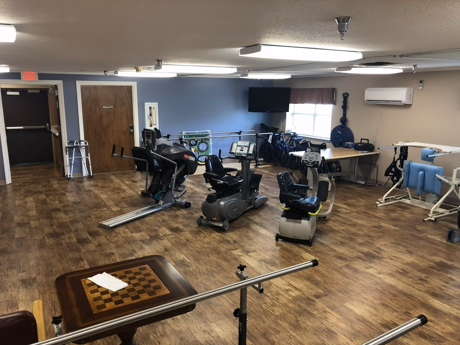 Rehabilitation exercise room with recumbent bikes, parallel bars, mobility aids and a wall-mounted TV.