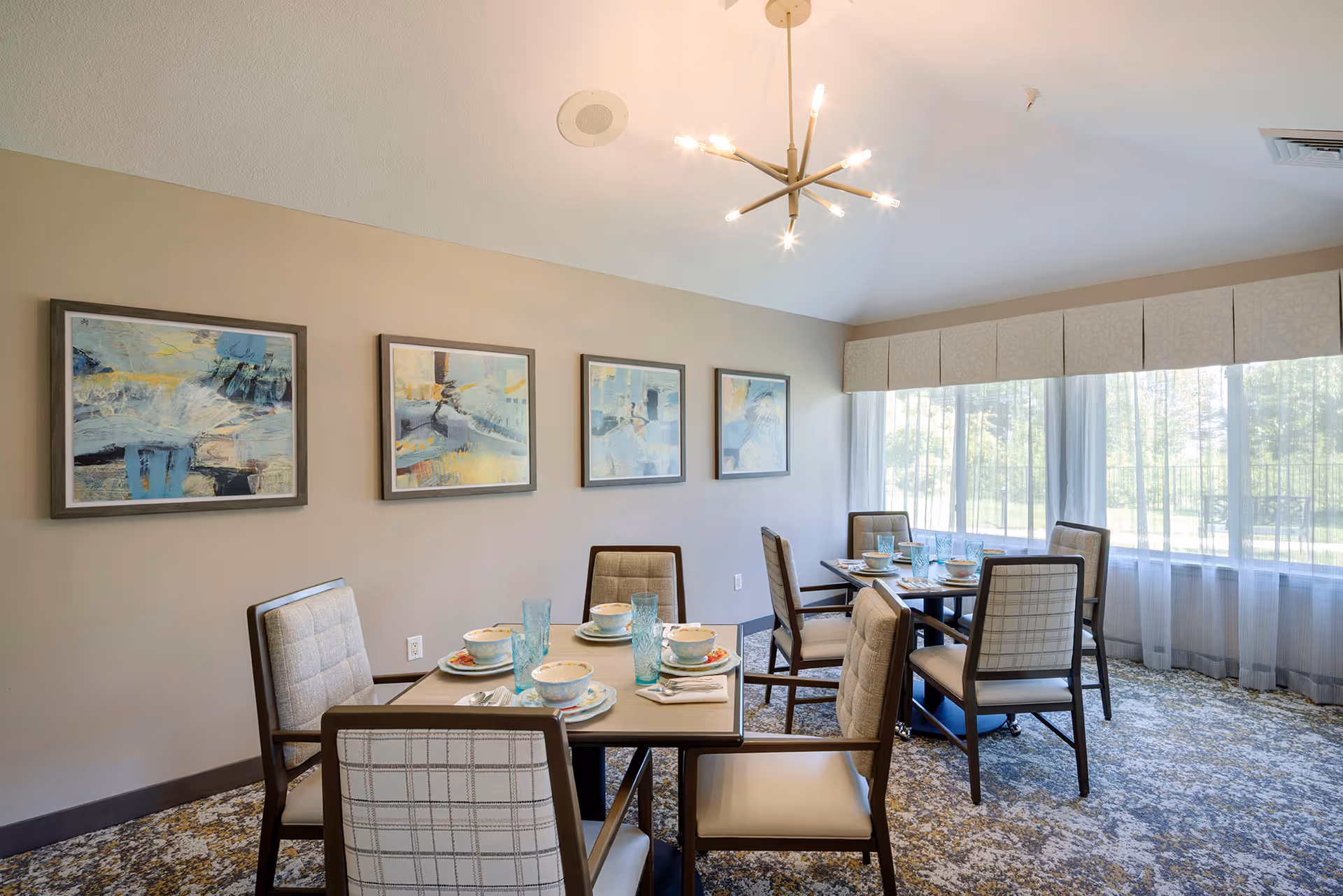 Bright dining room with multiple set tables, upholstered chairs, abstract wall art, and large windows with sheer curtains.