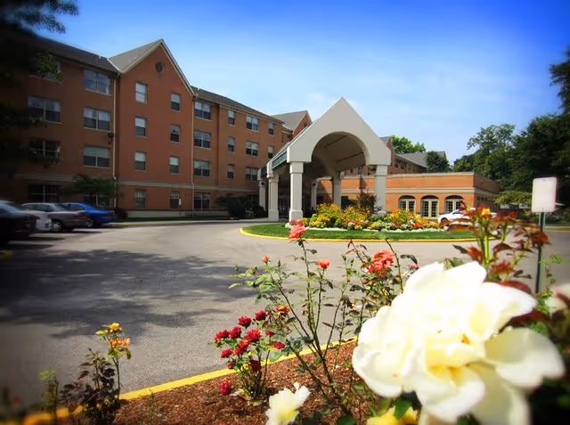Exterior view of a multi-story brick building with a covered entrance and a circular driveway. There are colorful flowers and bushes in the foreground and several parked cars along the side of the building under a clear blue sky.