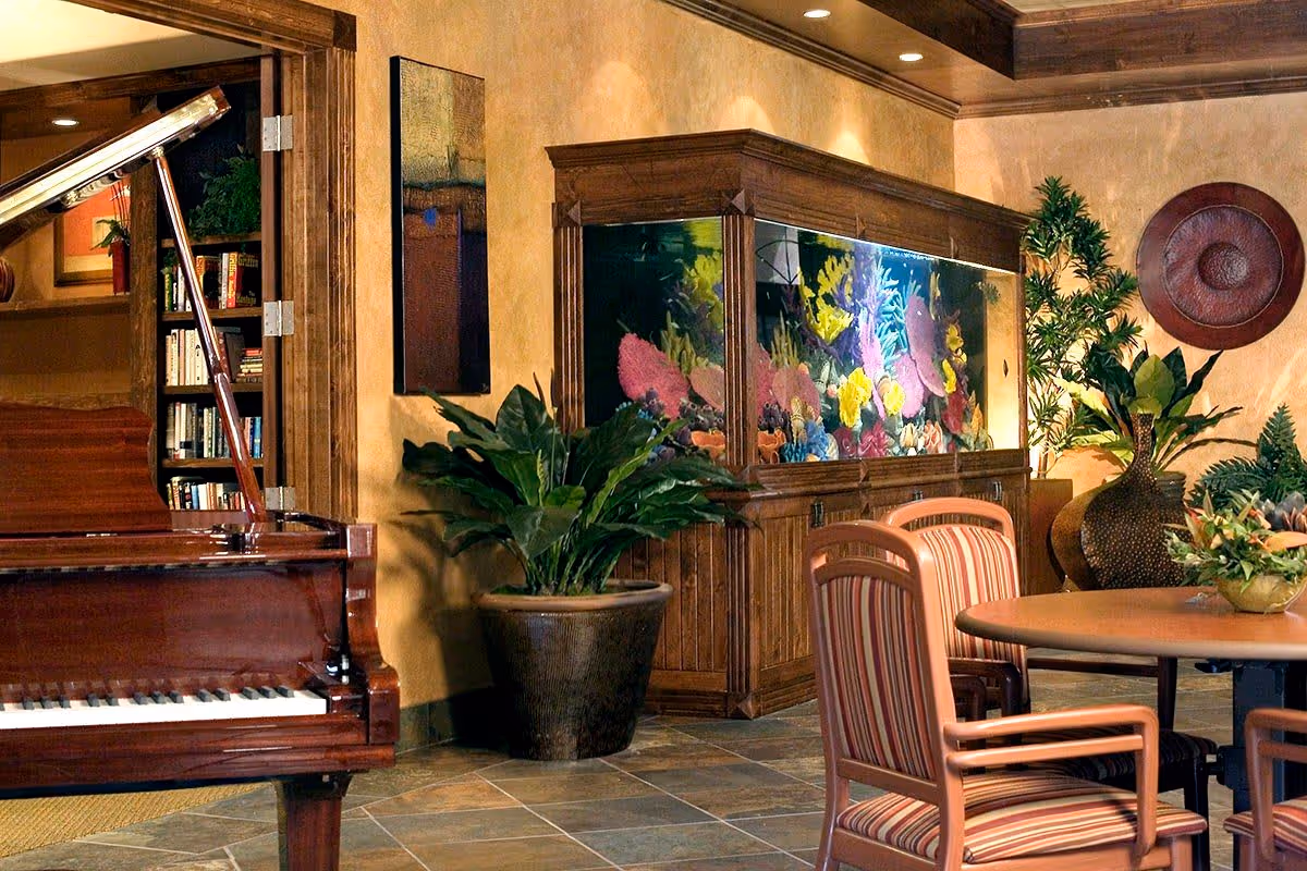 A cozy interior room featuring a grand piano on the left, a large built-in aquarium filled with colorful coral and fish in the center, and a round table with striped cushioned chairs on the right. The room is decorated with potted plants, a large decorative vase, and a wall-mounted circular art piece, with warm-toned walls and tiled flooring.