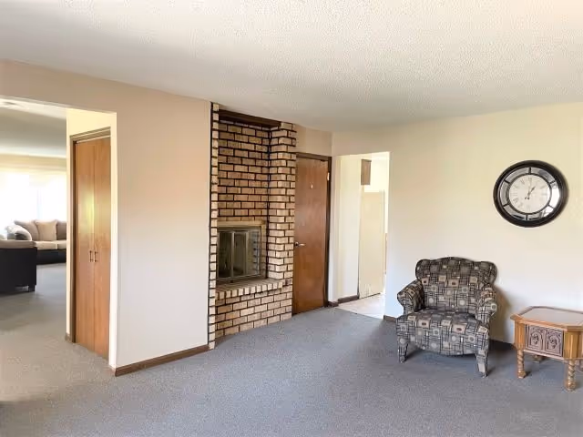 Interior view of a living area in an assisted living facility featuring a patterned armchair next to a small wooden side table with a drawer. A round wall clock hangs above the chair on a light-colored wall. There is a brick fireplace built into a partial wall, with a wooden door next to it. The floor is carpeted in gray, and an adjacent room with more seating is visible through an open doorway.