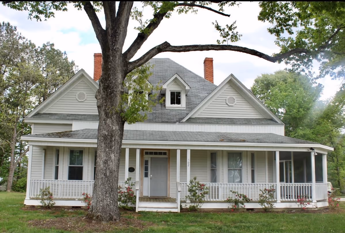 A two-story white house with a gray shingled roof, a front porch with white railings, and a large tree in the front yard. The house has multiple windows and two red brick chimneys. The porch extends to the right side where there is a screened-in section. The lawn is green with some small bushes and flowers near the porch.
