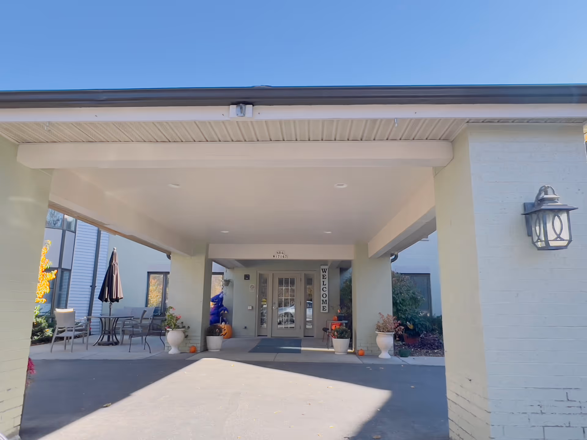 Covered main entrance and porte-cochère with a driveway, potted plants, outdoor seating, and a 'WELCOME' sign on the glass double doors.