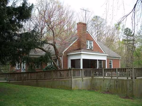 Red brick cottage with chimney, screened porch, and wooden fence in a grassy yard.