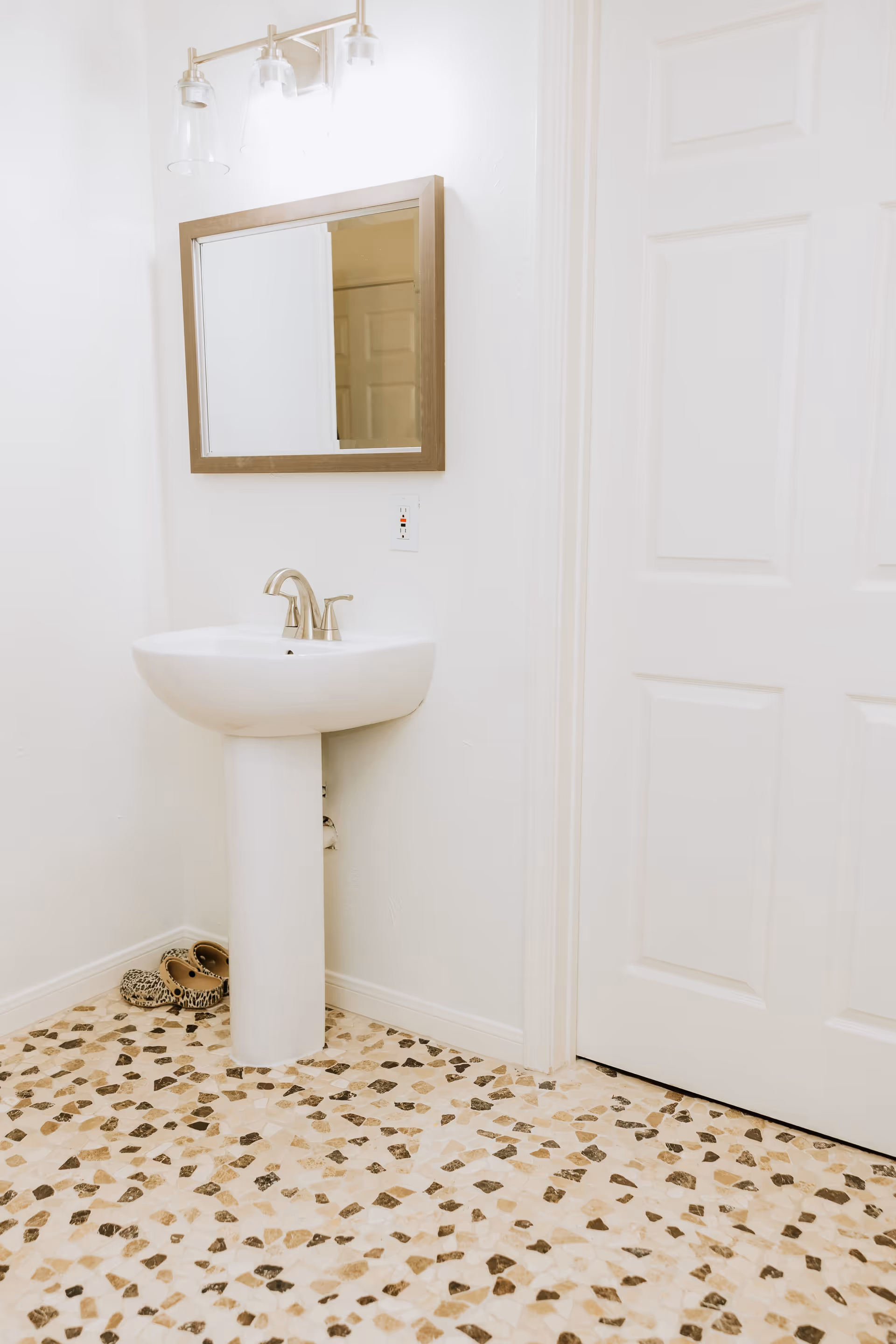 Small white bathroom with a pedestal sink, wall mirror and patterned tile floor next to a closed white door.
