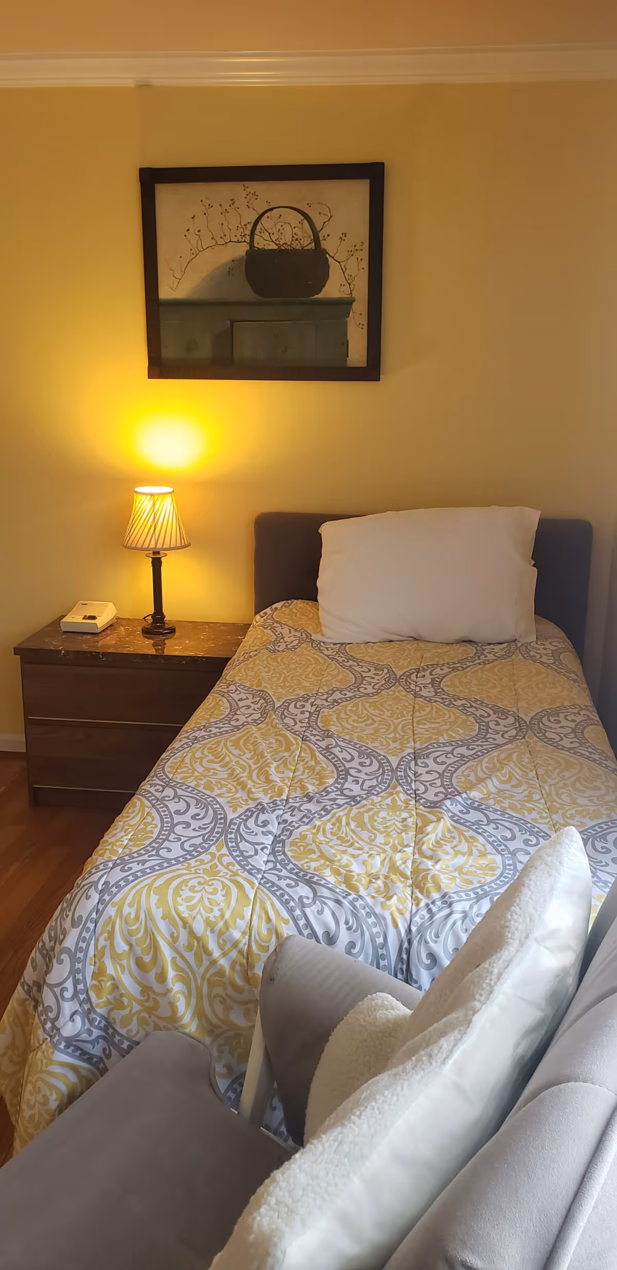 A single bed with yellow-and-gray patterned bedding next to a nightstand with a lamp and framed wall art in a small bedroom.