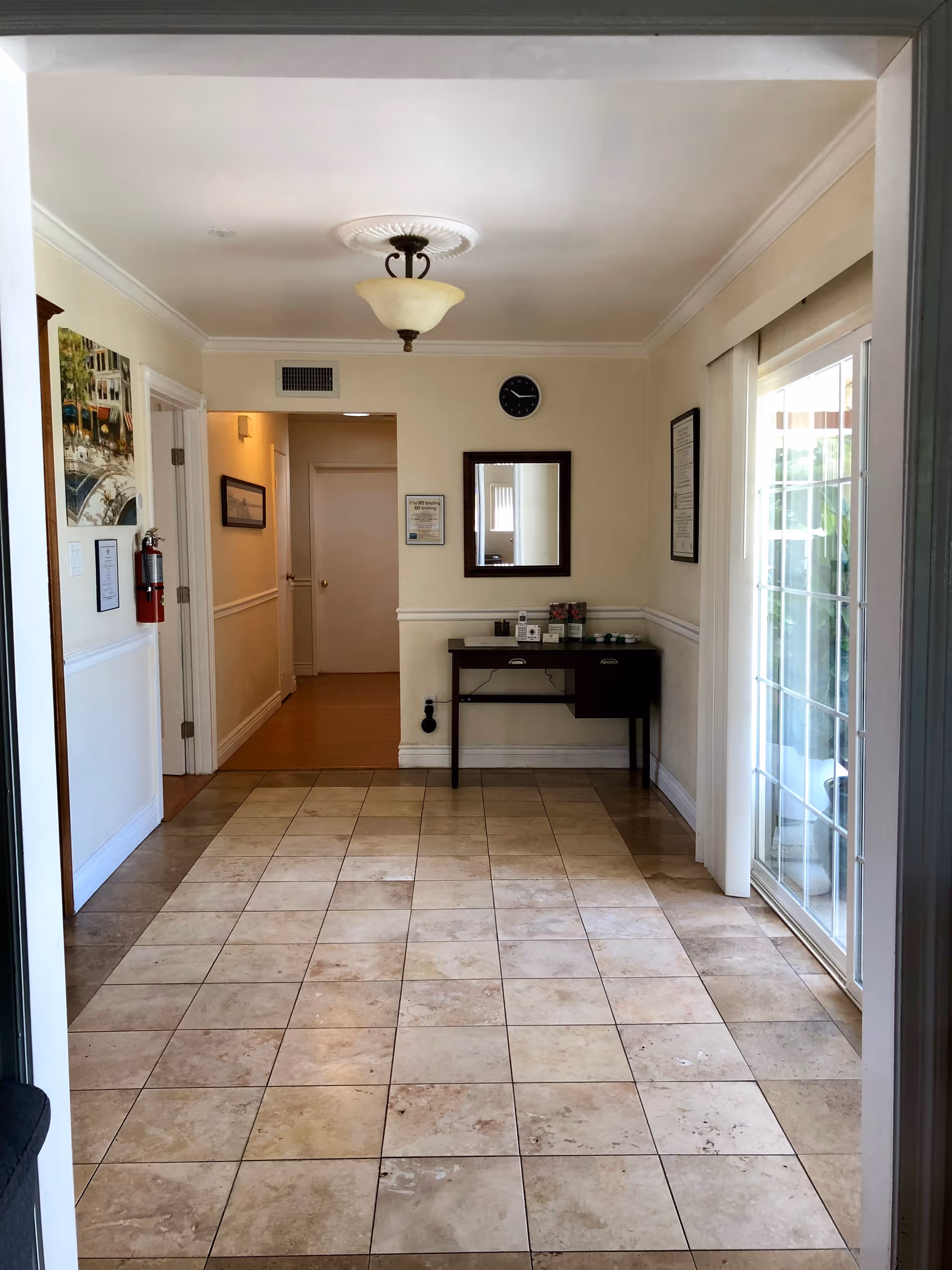 Interior hallway of Somerset Homecare with beige tiled floor, cream-colored walls, a ceiling light fixture, a wall clock, a mirror above a small dark wooden table with items on it, a fire extinguisher mounted on the left wall, framed pictures on the walls, and a sliding glass door on the right side leading outside.