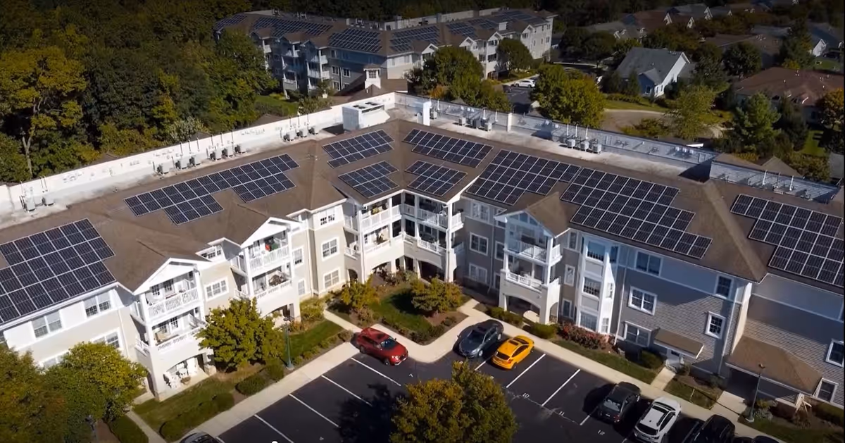 Aerial view of a multi-story senior living facility building with solar panels installed on the roof. The building is surrounded by trees and a parking lot with several cars parked. Residential houses and greenery are visible in the background.