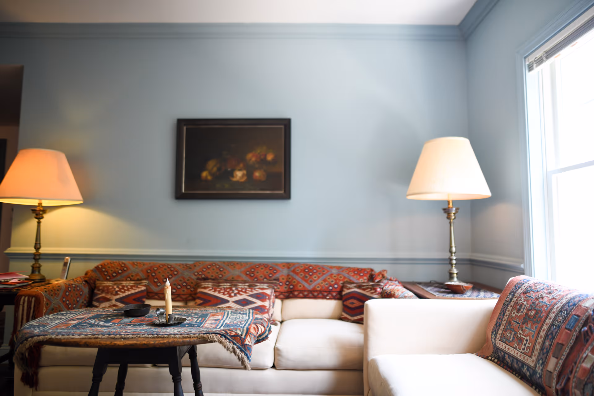 Cozy living room with a patterned sofa, a small coffee table with candles, two table lamps, and a framed painting on a pale blue wall.
