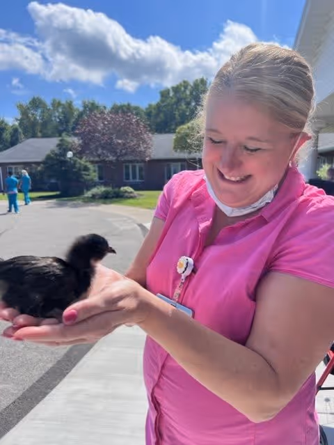 A smiling woman wearing a pink shirt and a face mask around her neck is holding a small black chick outdoors on a sunny day. In the background, there are trees, a building, and two people walking on a paved area.