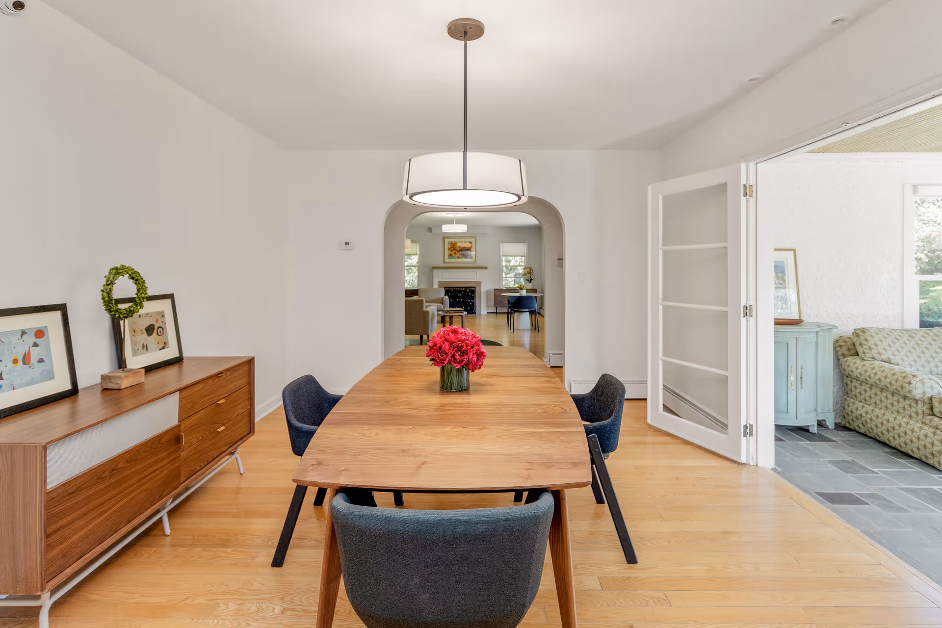 A modern dining room with a wooden table and four dark upholstered chairs. A vase with red flowers is centered on the table. On the left side, there is a wooden sideboard with framed artwork and a decorative wreath. The room has light wooden flooring and white walls. Through an arched doorway, a living room with a fireplace and additional seating is visible. To the right, glass-paneled double doors open to a sunroom with a patterned sofa and a small blue cabinet.