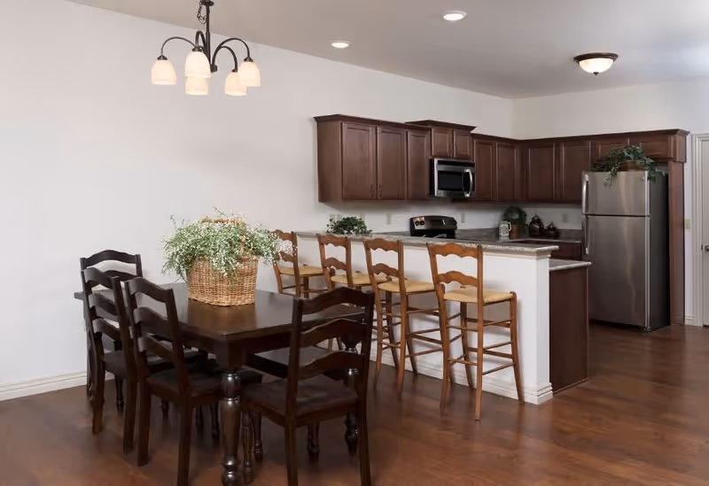 Open-plan kitchen and dining area with a wooden dining table, four bar stools at a counter, dark wood cabinets and stainless steel appliances.
