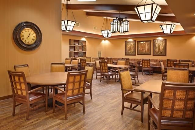 A spacious dining room with multiple wooden tables and chairs arranged neatly on a wood floor. The walls are decorated with a large round clock and framed artwork. The ceiling features several hanging lantern-style light fixtures providing warm lighting.