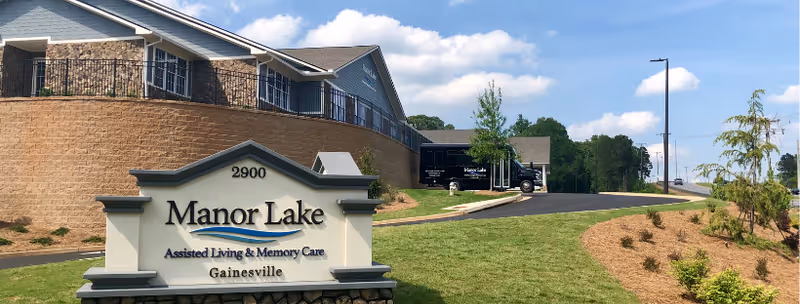 Exterior view of Manor Lake Assisted Living & Memory Care in Gainesville showing a large sign with the facility name in front of a two-story building with stone and siding exterior, a driveway with a black truck parked, and landscaped grassy areas with small trees and shrubs under a partly cloudy sky.