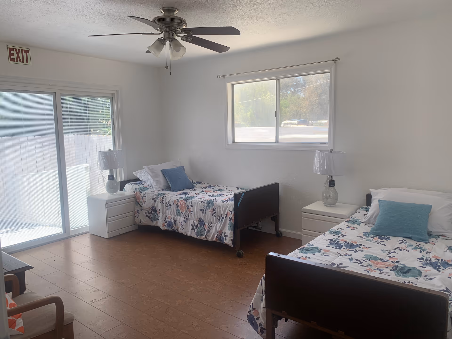 Bright shared bedroom with two single beds with floral bedding, bedside tables and lamps, a ceiling fan, and a sliding glass door.